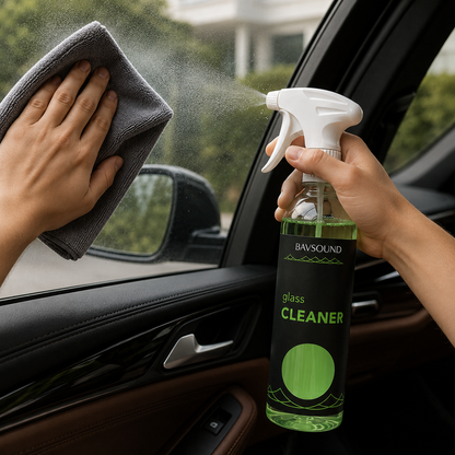 A person holds a spray bottle of BAVSOUND glass cleaner and sprays a car window while wiping it with a gray microfiber cloth. The product is shown in a real-world car interior setting.